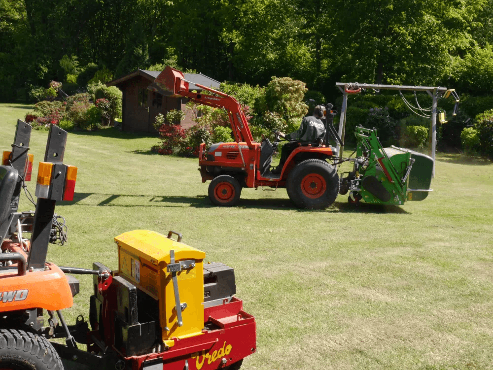Ein Traktor mit einem Anhänger und einer grünen Maschine zur Rasenpflege fährt über das gepflegte Gras, während eine weitere Maschine im Vordergrund zu sehen ist. Der Garten im Hintergrund ist von Bäumen und Sträuchern umgeben.