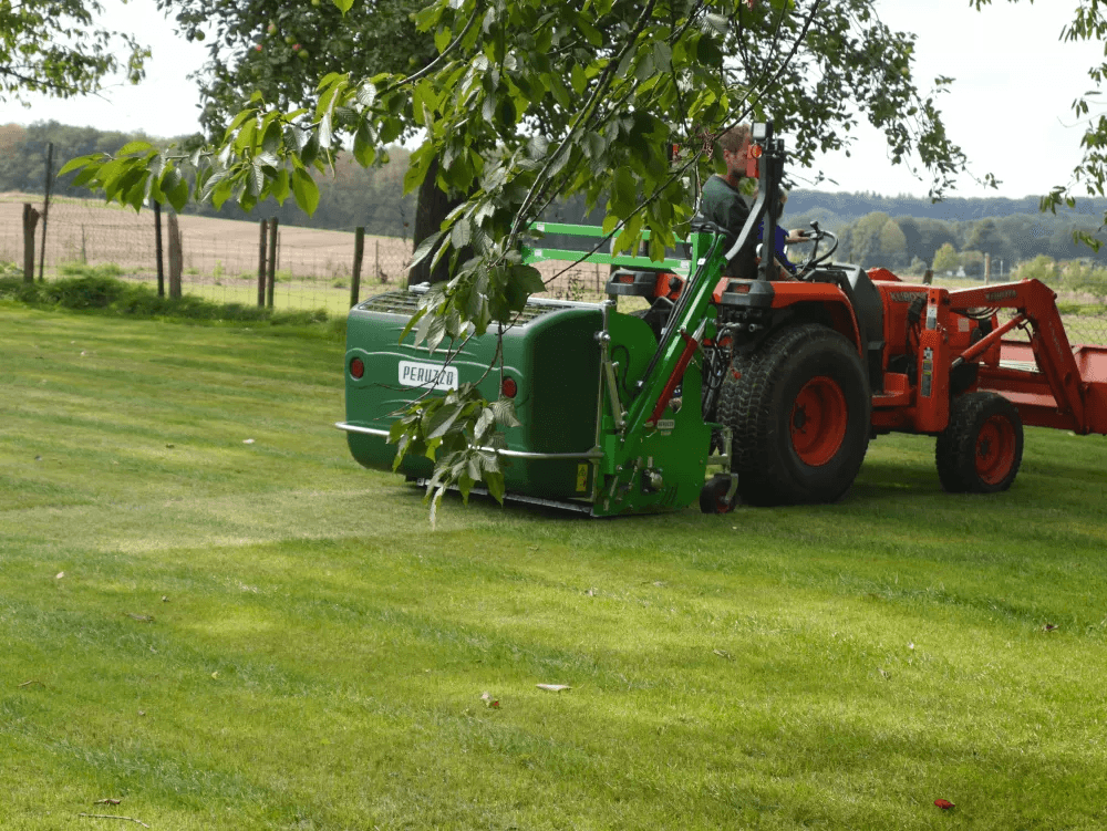 Ein Traktor mit einer grünen Maschine zur Rasenpflege fährt über den gepflegten Rasen, während im Hintergrund ein weites, offenes Feld sichtbar ist. Der Traktor ist von einem Baum im Vordergrund umrahmt.
