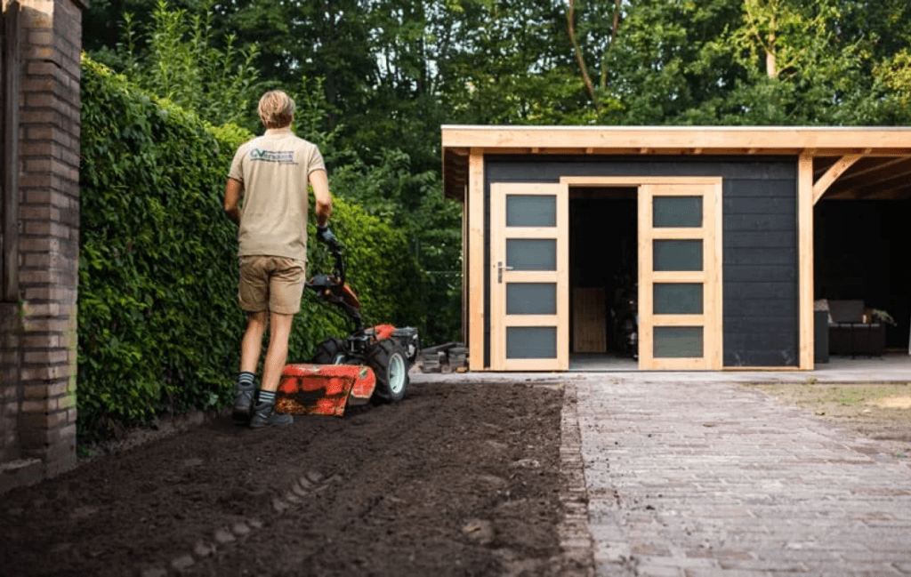 Ein Gärtner arbeitet mit einer Bodenfräse, um den Boden für die Rasenverlegung vorzubereiten. Der Gartenbereich ist sauber und gut bearbeitet, während im Hintergrund ein Gartenhaus zu sehen ist.