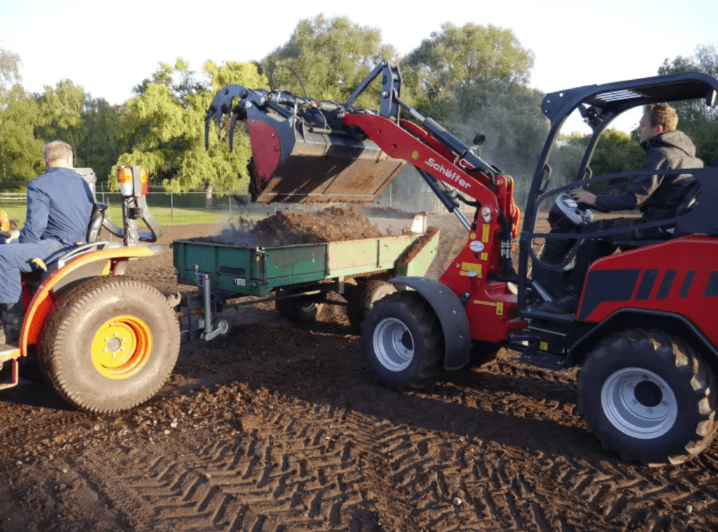 Ein Schaufellader lädt Erde in einen Anhänger. Der Fahrer steuert die Maschine, während der Arm des Laders Erde auflädt.
