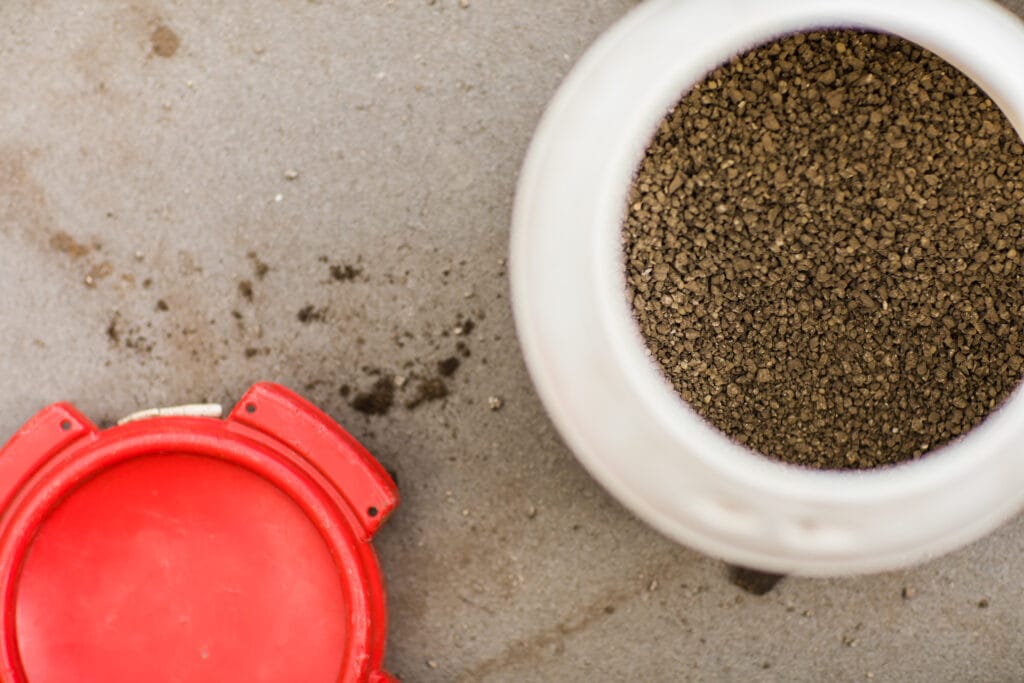 Ein Eimer mit Rasendünger, der auf dem Boden steht. Der Dünger besteht aus kleinen, gleichmäßigen Granulen und ist ideal für die Pflege von Rollrasen, um das Wachstum und die Gesundheit des Grases zu fördern.