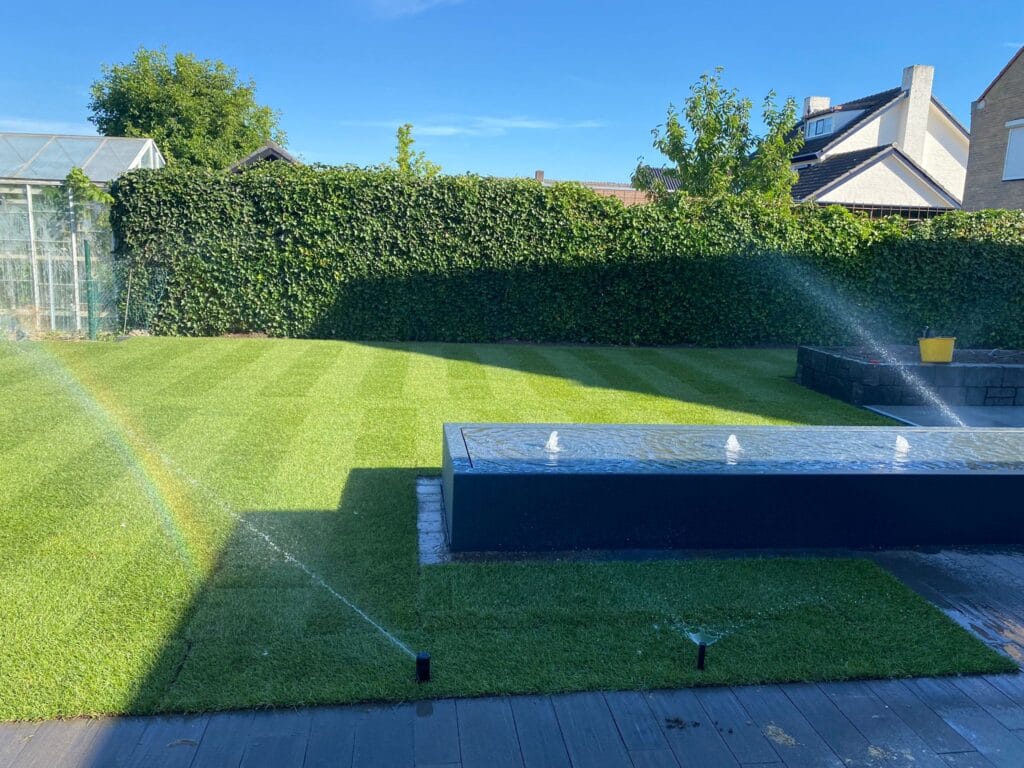 Ein grüner, gut gepflegter Garten mit einem bewässerten Rasen und einem Springbrunnen. Im Hintergrund sprüht ein Bewässerungssystem Wasser auf das Gras, während der Rasen gleichmäßig gepflegt und das Wetter klar ist.