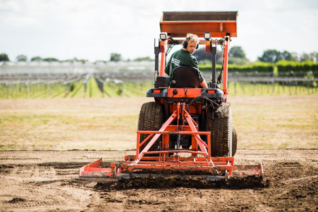 Ein Rasenprofi fährt einen Traktor mit einer Bodenbearbeitungsmaschine über ein Feld, um den Boden für die Aussaat vorzubereiten. Im Hintergrund sind Felder und Gewächshäuser zu sehen.