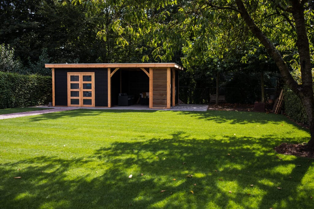 Ein gepflegter Garten mit einem frisch nachgesäten Rasen. Im Hintergrund steht ein Gartenhaus, das von Bäumen und Sträuchern umgeben ist. Der Rasen zeigt eine gleichmäßige grüne Farbe.