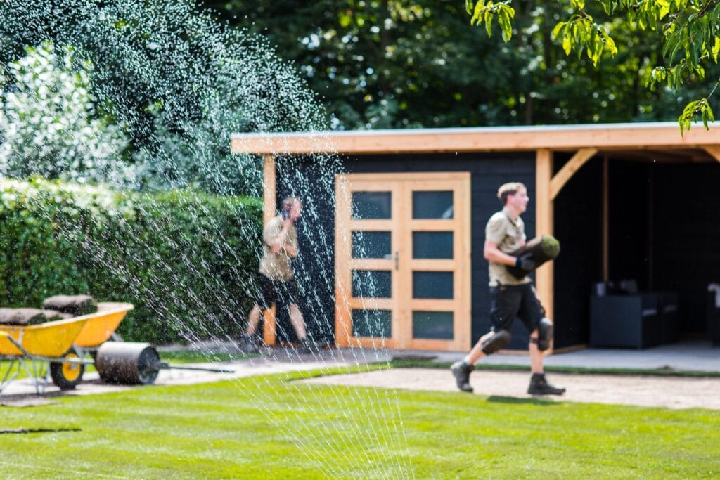 Nahaufnahme eines laufenden Rasensprengers, während im Hintergrund zwei Gärtner Rollrasen verlegen und eine gelbe Schubkarre mit Rasensoden steht.